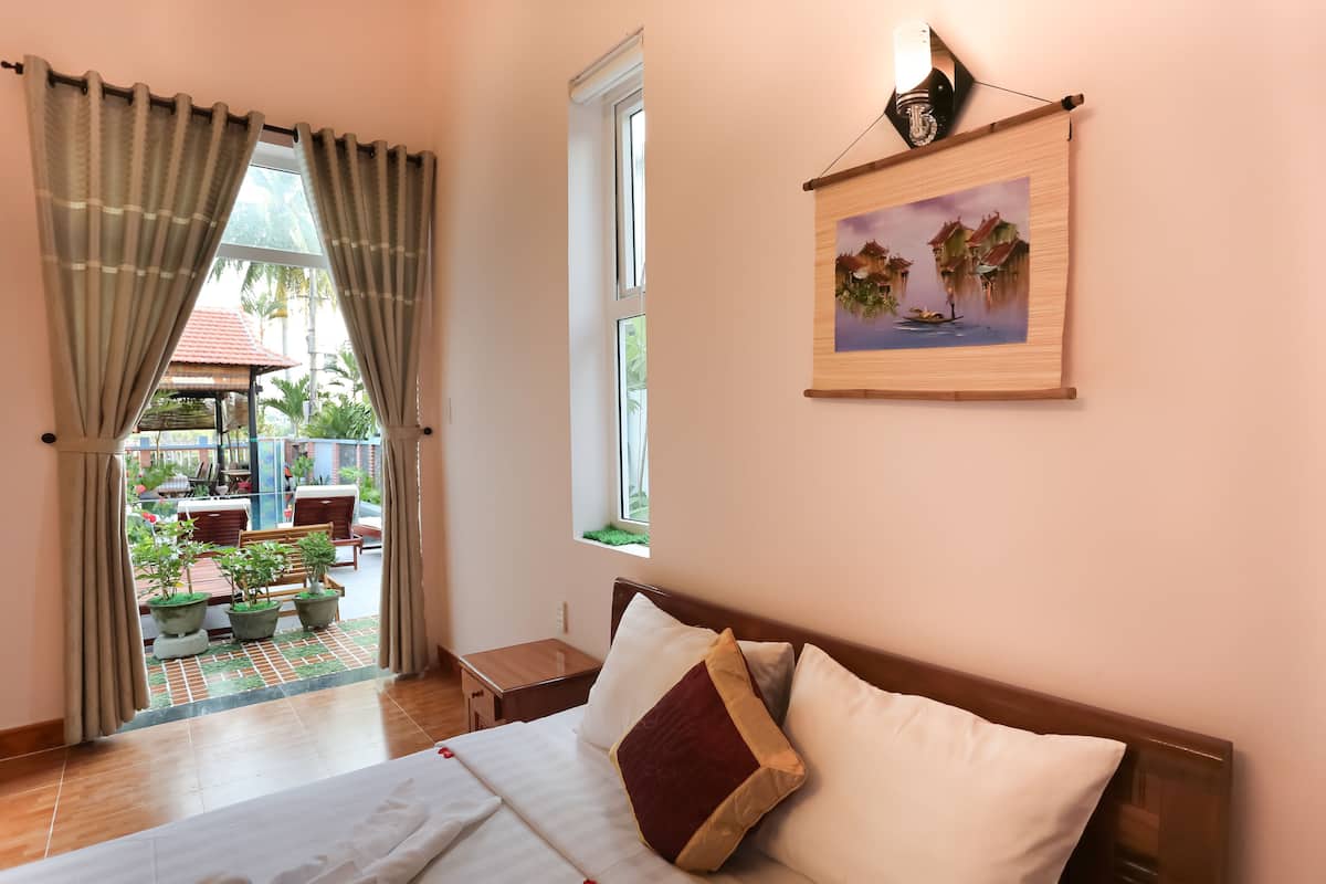 Une chambre d’hôtel comprenant un lit, une petite table de chevet, une fenêtre avec des rideaux, un tableau accroché au mur et une vue sur un espace extérieur agrémenté de plantes et de mobilier.