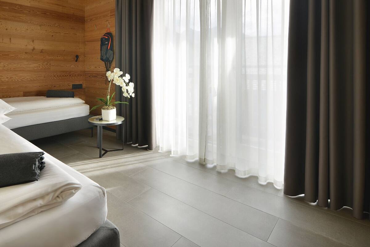 A modern hotel room with two beds, a small table with a potted plant, and a view of the outdoors through sheer curtains.