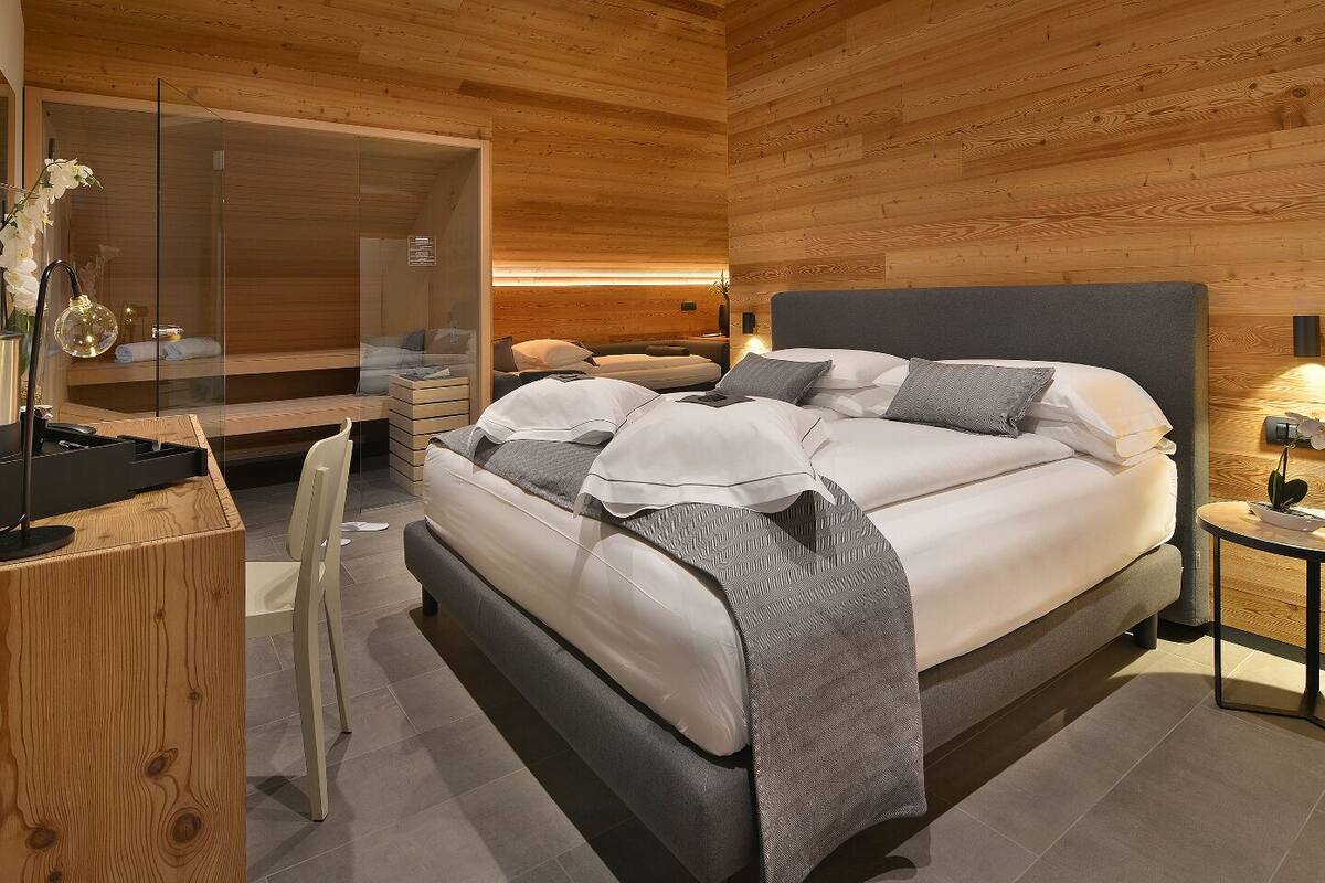 A modern bedroom with a wooden ceiling, a large bed with grey bedding, a bedside table with a lamp, and a desk with a computer and a coffee machine.