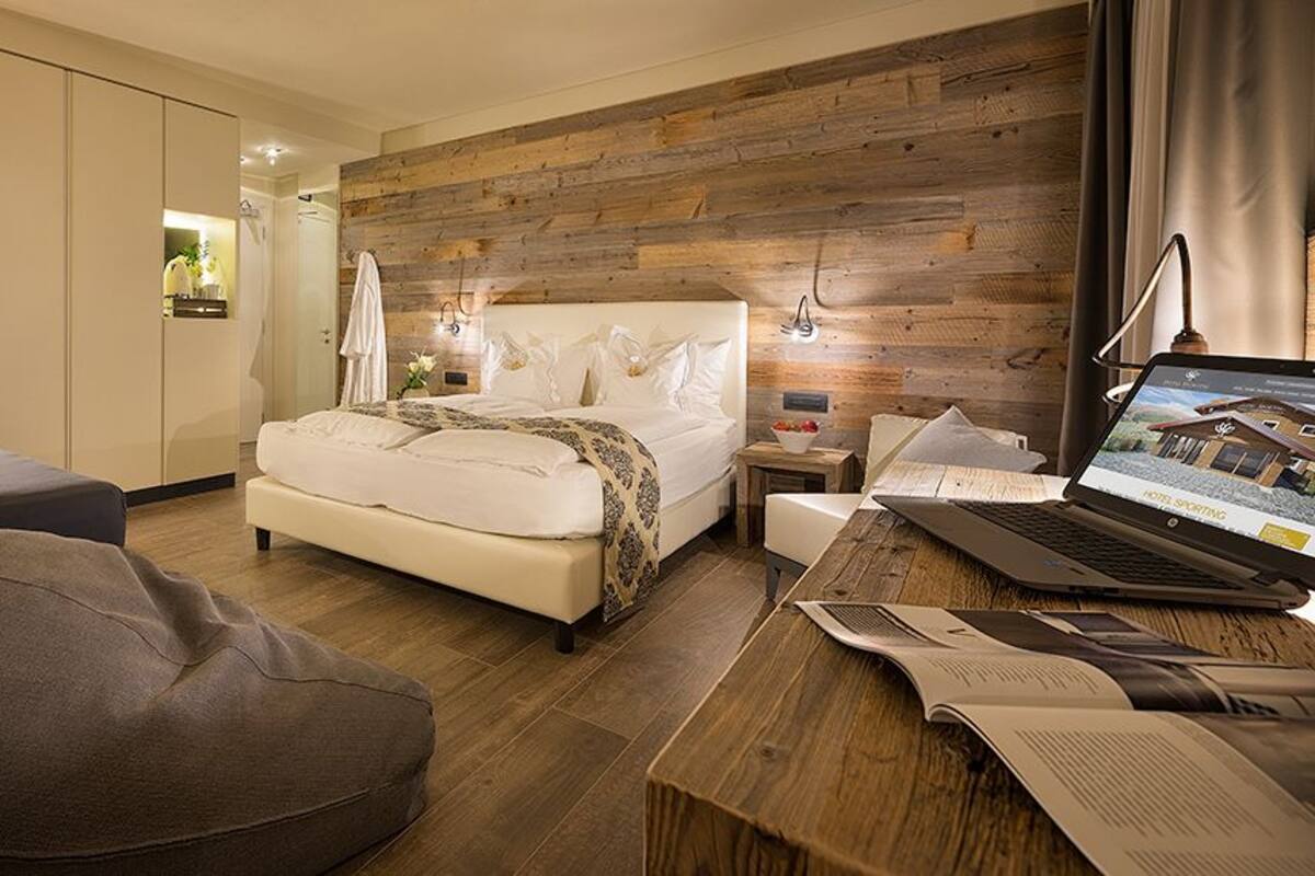 A modern hotel room with a bed, a desk with a laptop, a sofa, and a wooden accent wall.