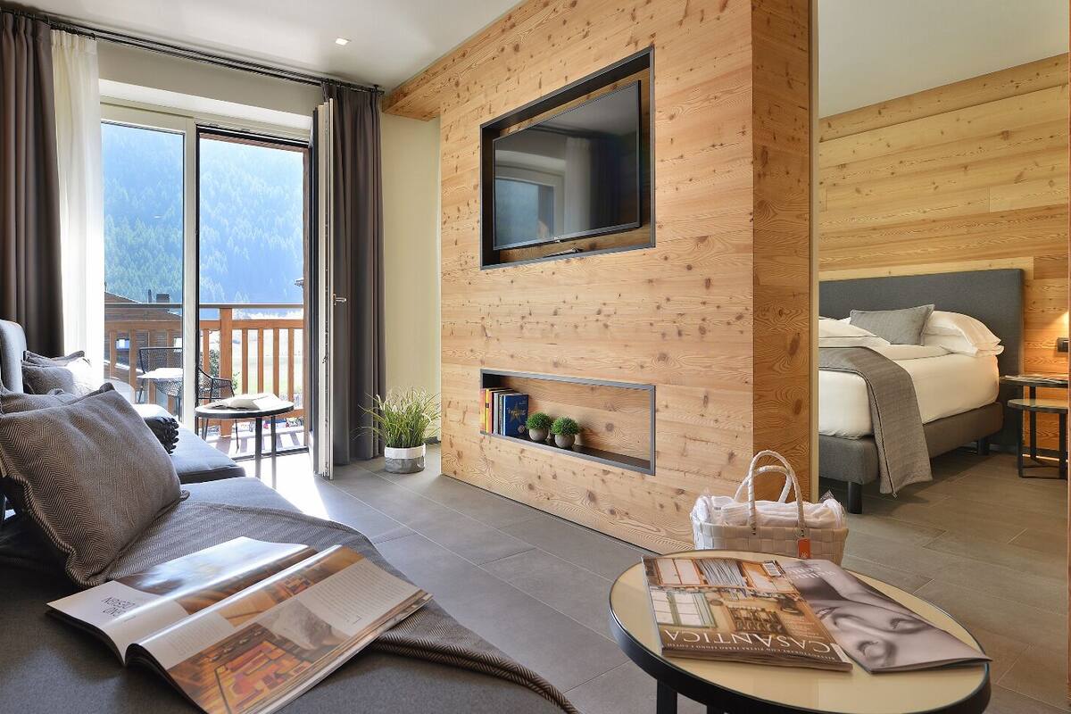 A modern hotel room with a sofa, a small table with a magazine, a TV mounted on a wooden wall, a bed with a headboard, and a balcony with a view of the mountains.