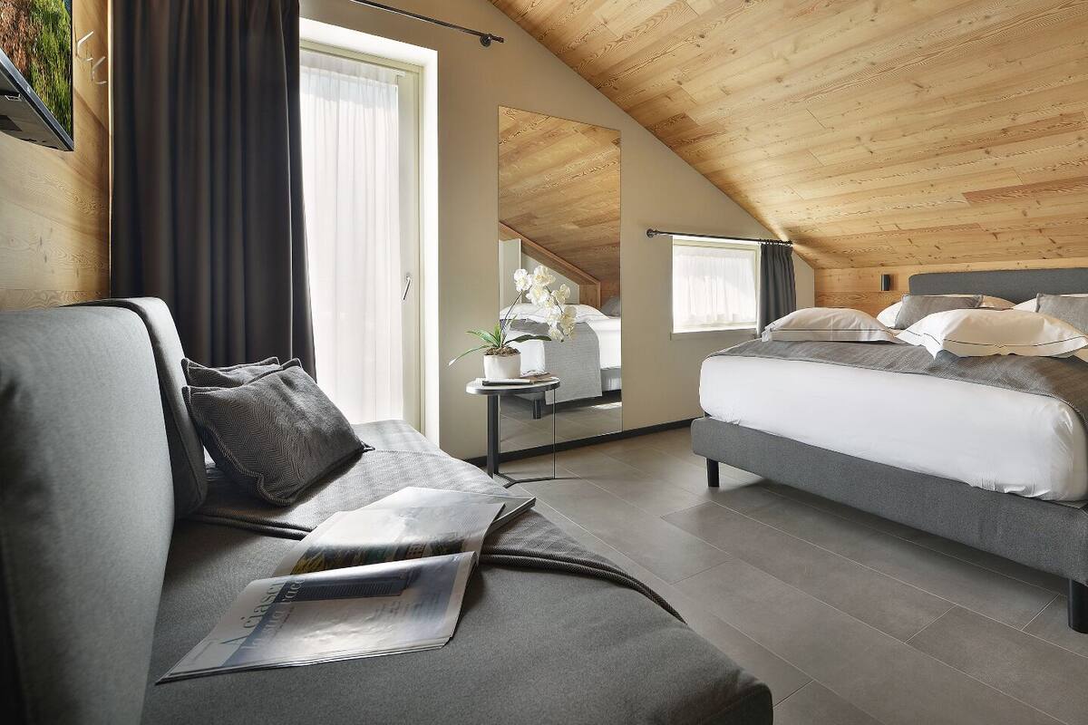 A modern bedroom with a sloped ceiling, a large bed with grey bedding, a small bedside table, and a chair with a cushion.