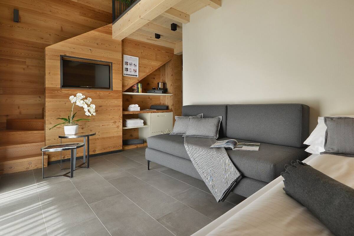A modern hotel room with a wooden interior, a grey sofa, a bed with grey and white bedding, a small table with a plant, and a TV mounted on the wall.