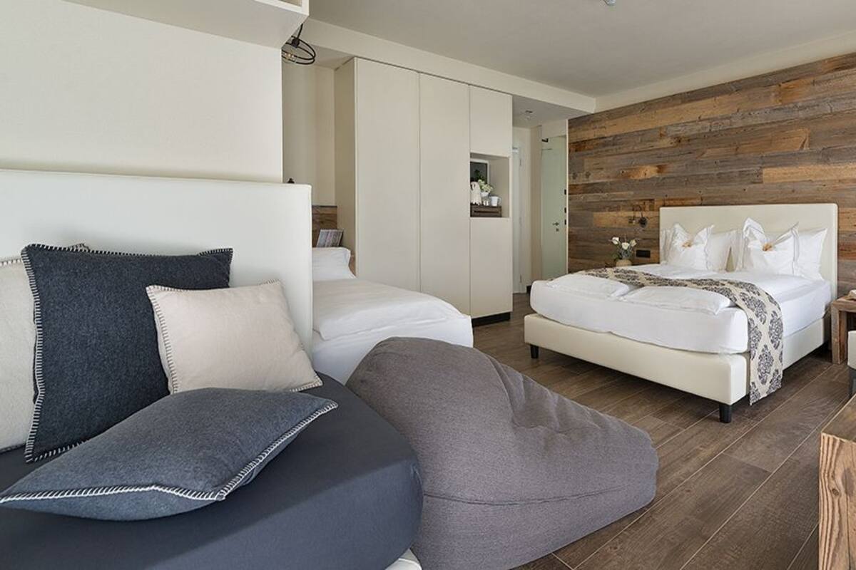 A modern hotel room with a wooden accent wall, a large bed, a sofa, a desk with a laptop, and a small bedside table.