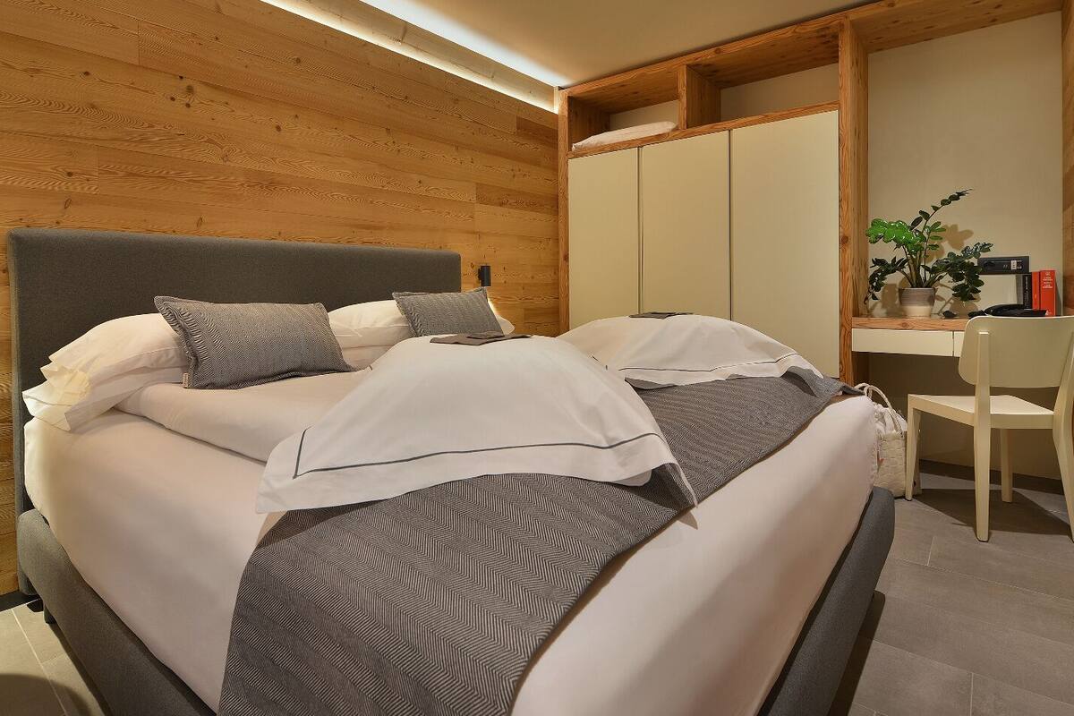 A modern bedroom with a wooden ceiling, a bed with white and gray bedding, a bedside table with a lamp, and a desk with a chair.