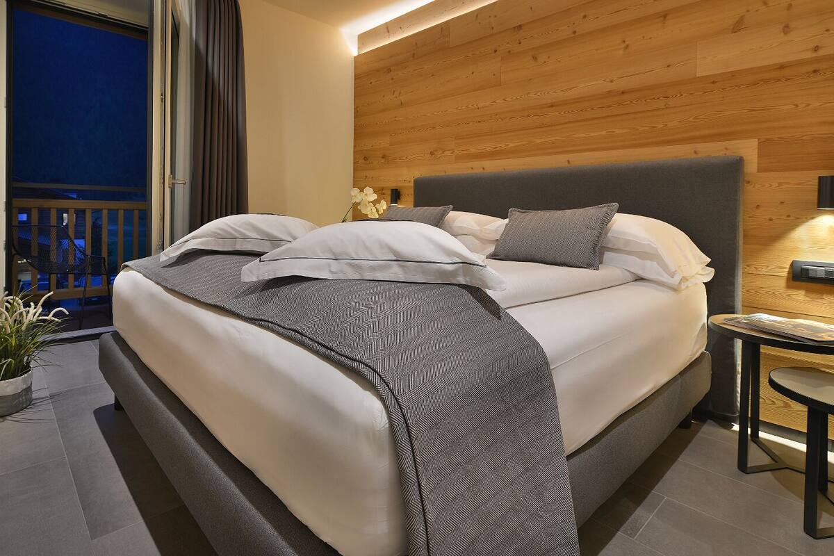 A modern bedroom with a large bed, a small bedside table, a potted plant, and a view of the balcony.