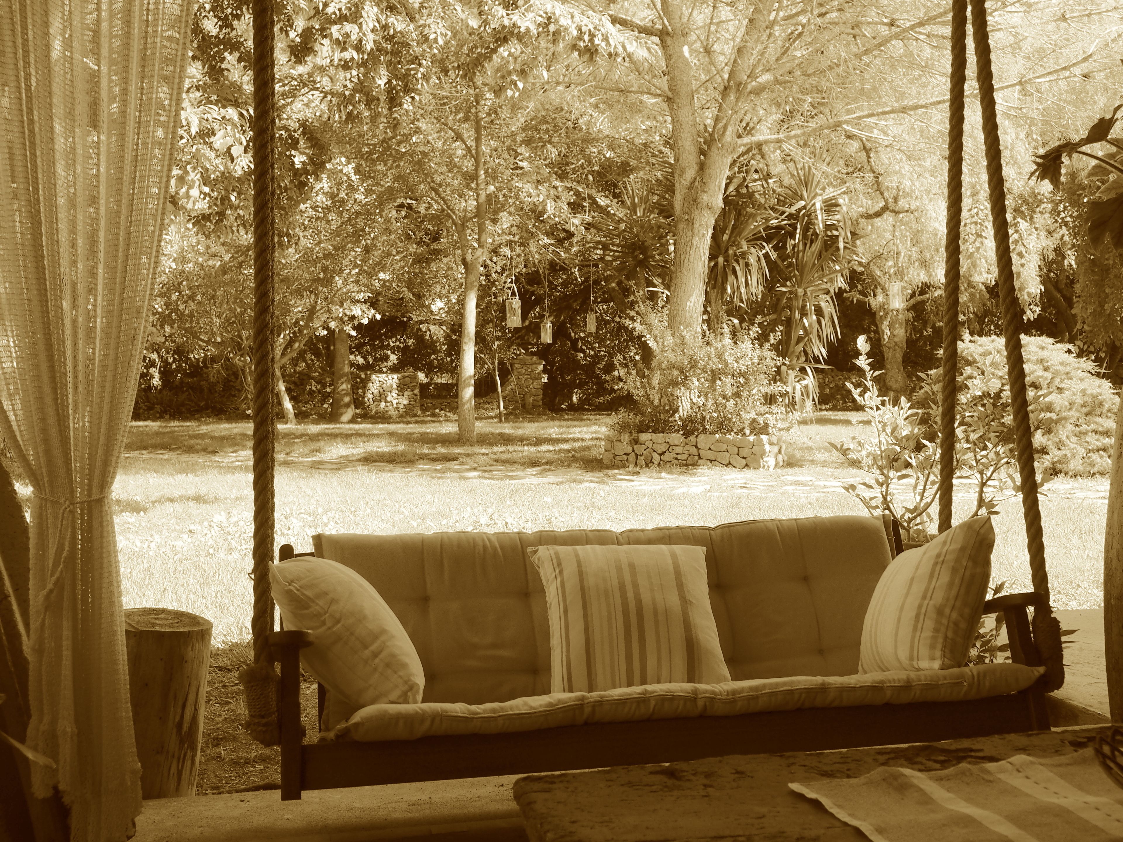 Un banc en bois avec des coussins, situé dans un espace jardin agrémenté d’arbres et de plantes.