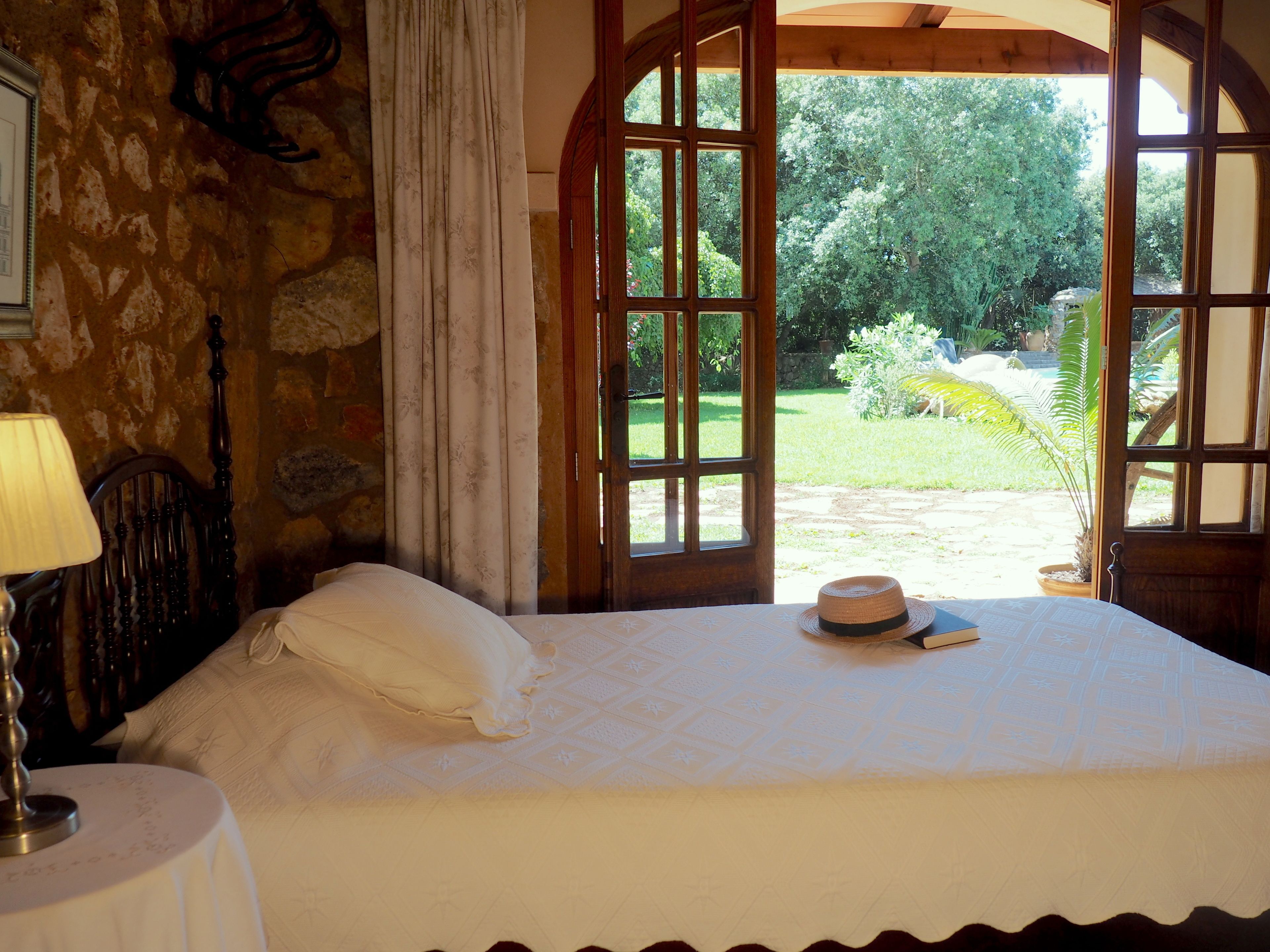A bedroom with a bed, a lamp, and a view of a garden through open windows.