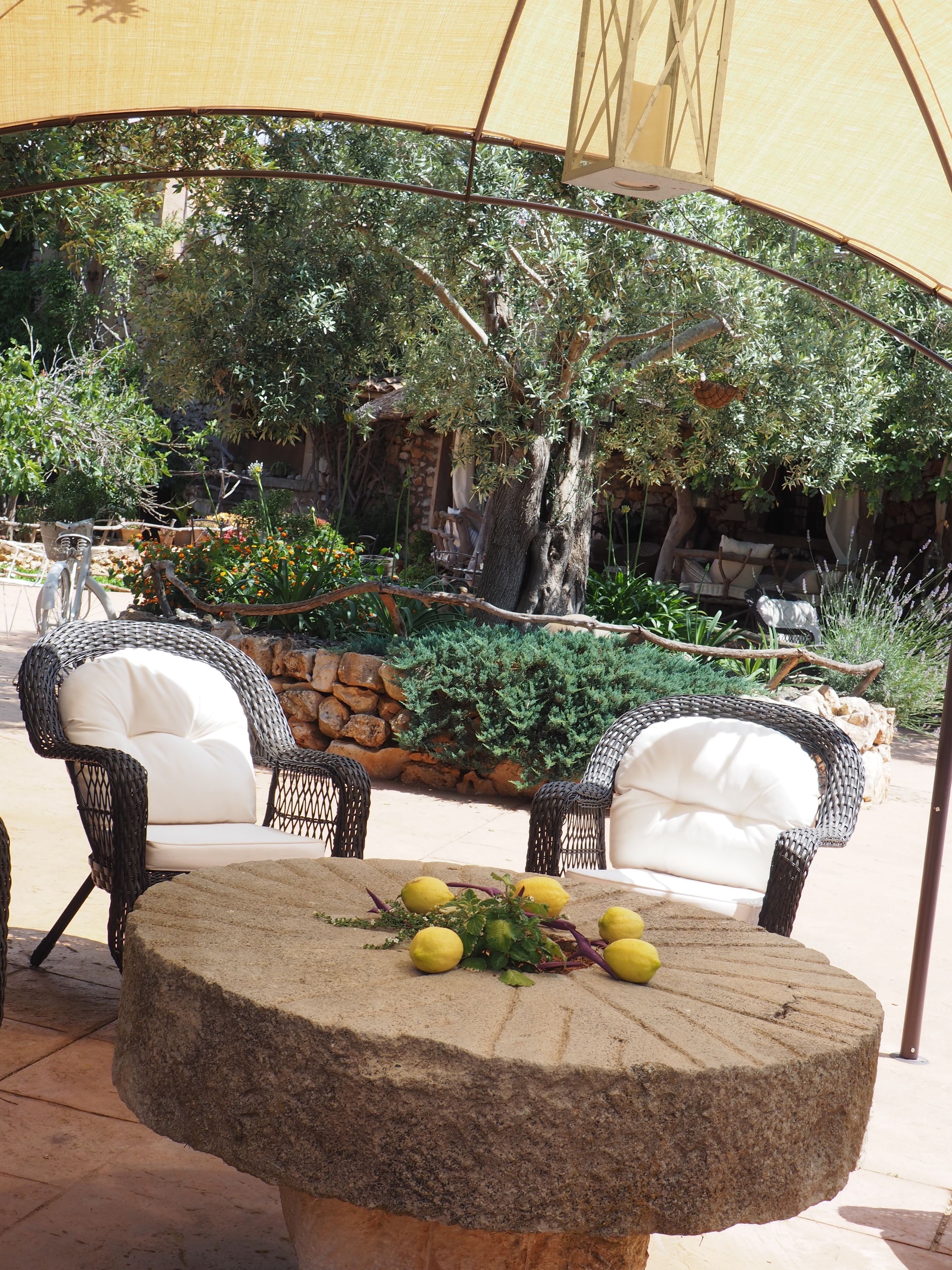 An outdoor seating area with wicker chairs and a stone table, surrounded by greenery and a stone wall.