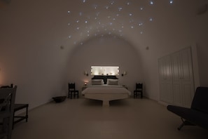 A bedroom with a bed centered under a curved ceiling, flanked by two nightstands with lamps, a wardrobe, and a chair. - Karpimo Horizon - Caldera View with private hot tub (Santorini)