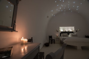 Un dormitorio con poca luz, que incluye una cama, una mesita de noche y un espejo. Hay velas sobre la mesa y un candelabro en el techo.