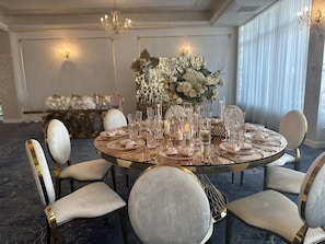 Una mesa redonda con superficie de vidrio, rodeada por sillas blancas tapizadas con detalles dorados, preparada para un evento formal.