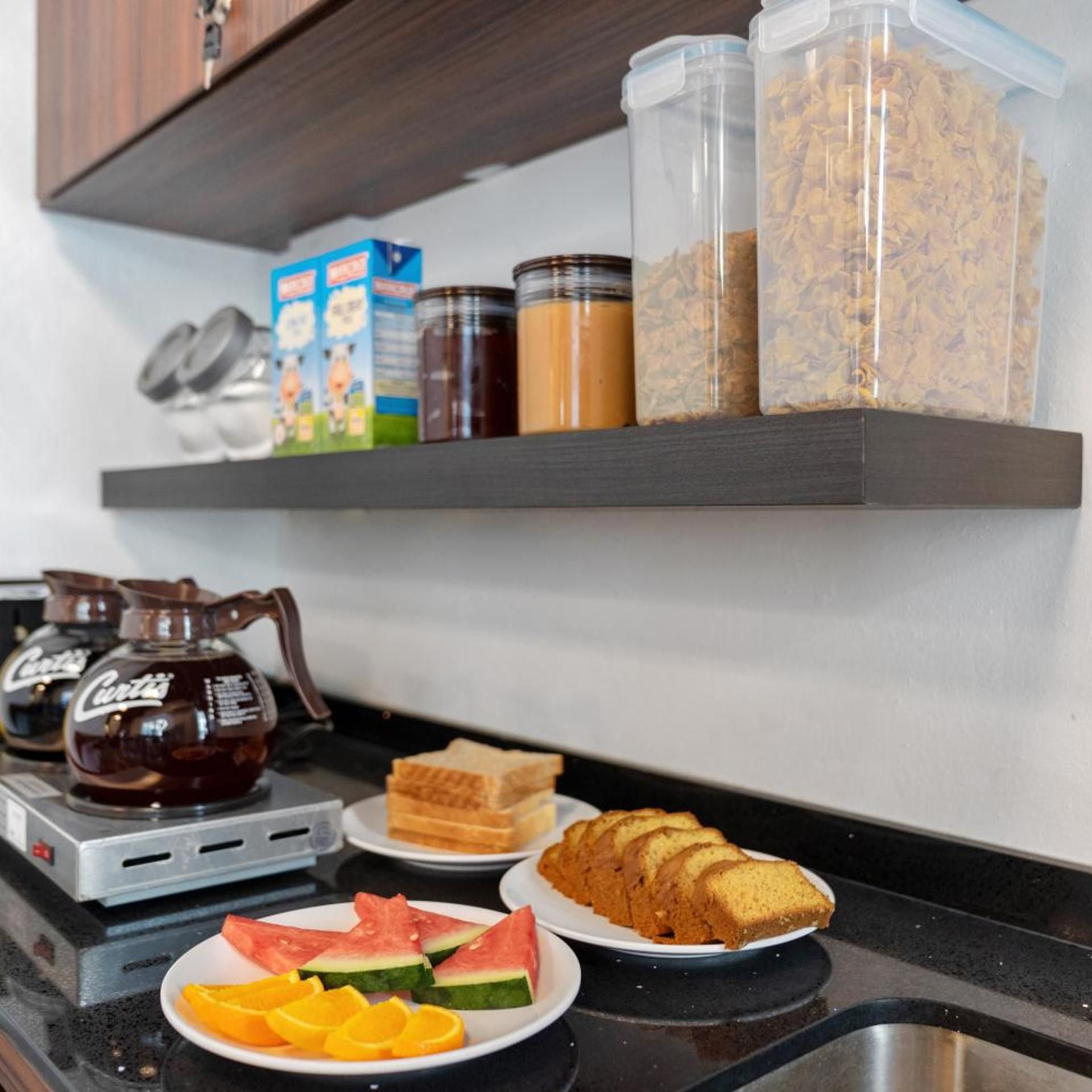 Una encimera de cocina con una cafetera, cajas de cereales, frascos de condimentos y platos de comida que incluyen pan y fruta.