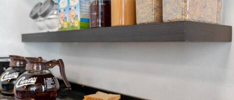 Una encimera de cocina con una cafetera, cajas de cereales, frascos de condimentos y platos de comida que incluyen pan y fruta.