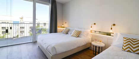 Habitación de hotel con una cama grande, una mesita de noche pequeña, una silla y un balcón con vistas a edificios.