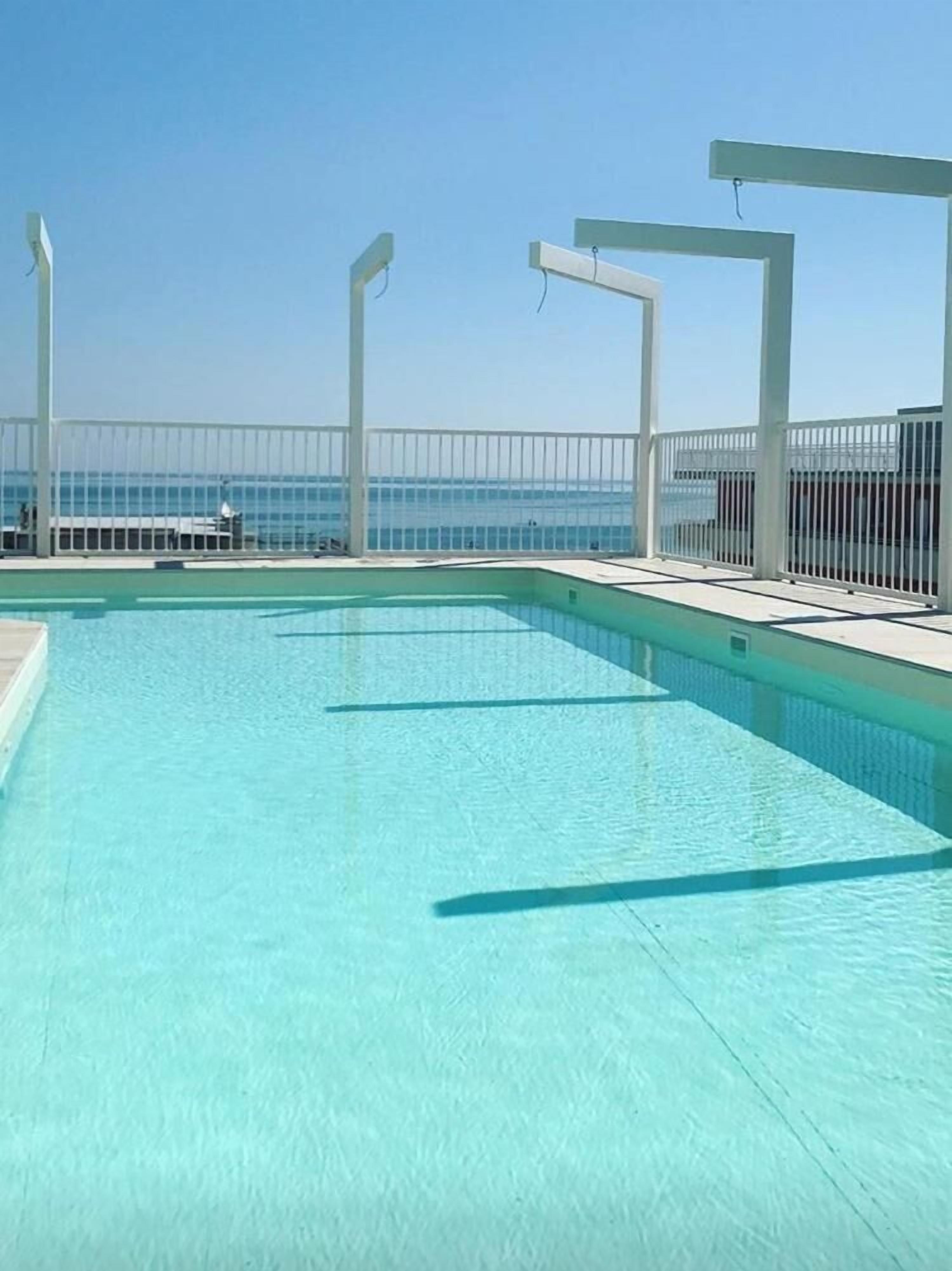 Ein klares, blaues Schwimmbecken mit einer weißen Terrasse und einem weißen Zaun mit Blick auf den Ozean.