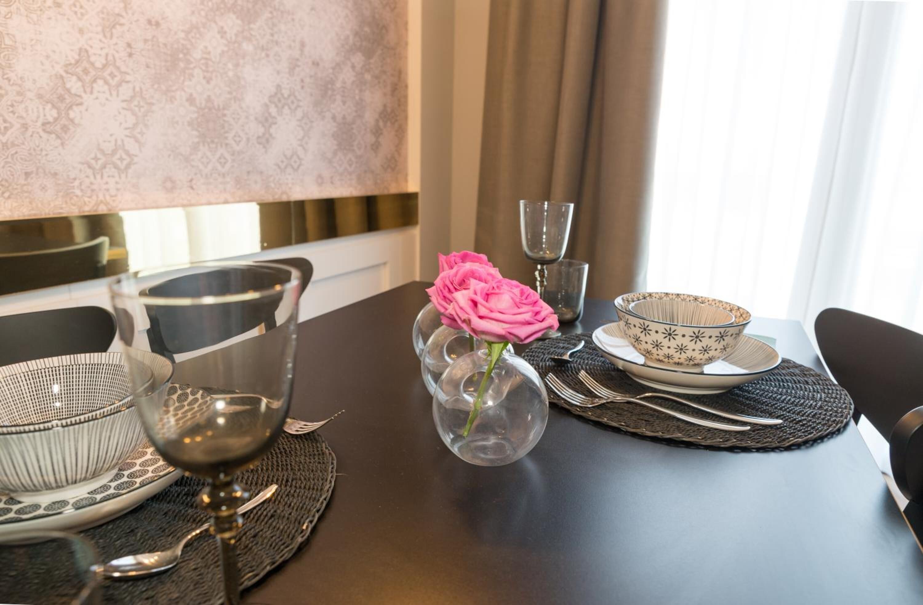 Une table à manger est dressée avec un vase en verre transparent contenant une rose rose, un bol à motifs et un verre à vin.