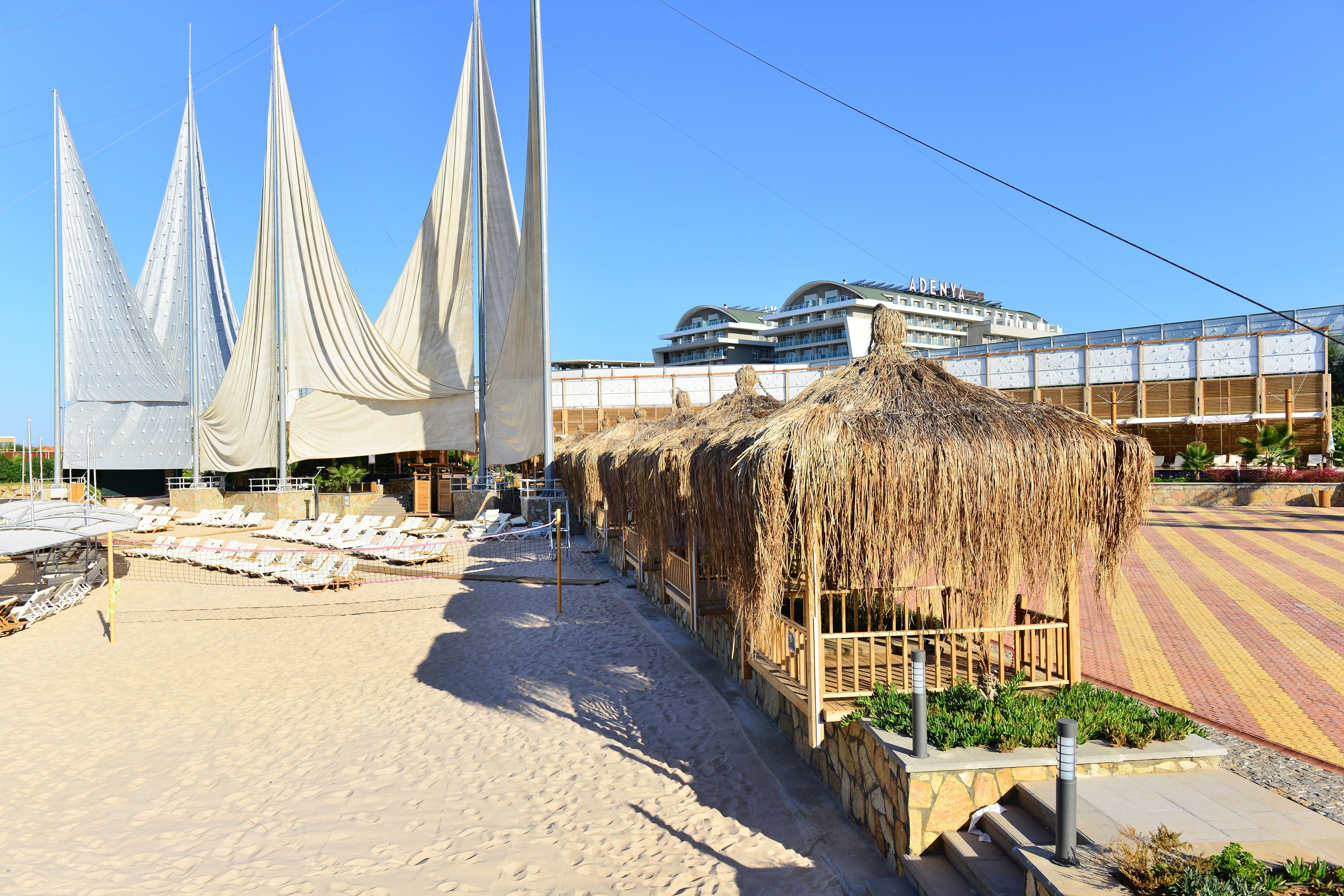 Un hotel frente a la playa con estructuras de techo de paja, un área de piscina y un edificio moderno al fondo.