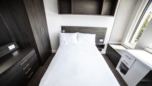 A modern hotel room with a large bed, dark wooden wardrobe, a small desk, and a window providing a view of the outdoors. - University of Bath Guest Accommodation (Bath)