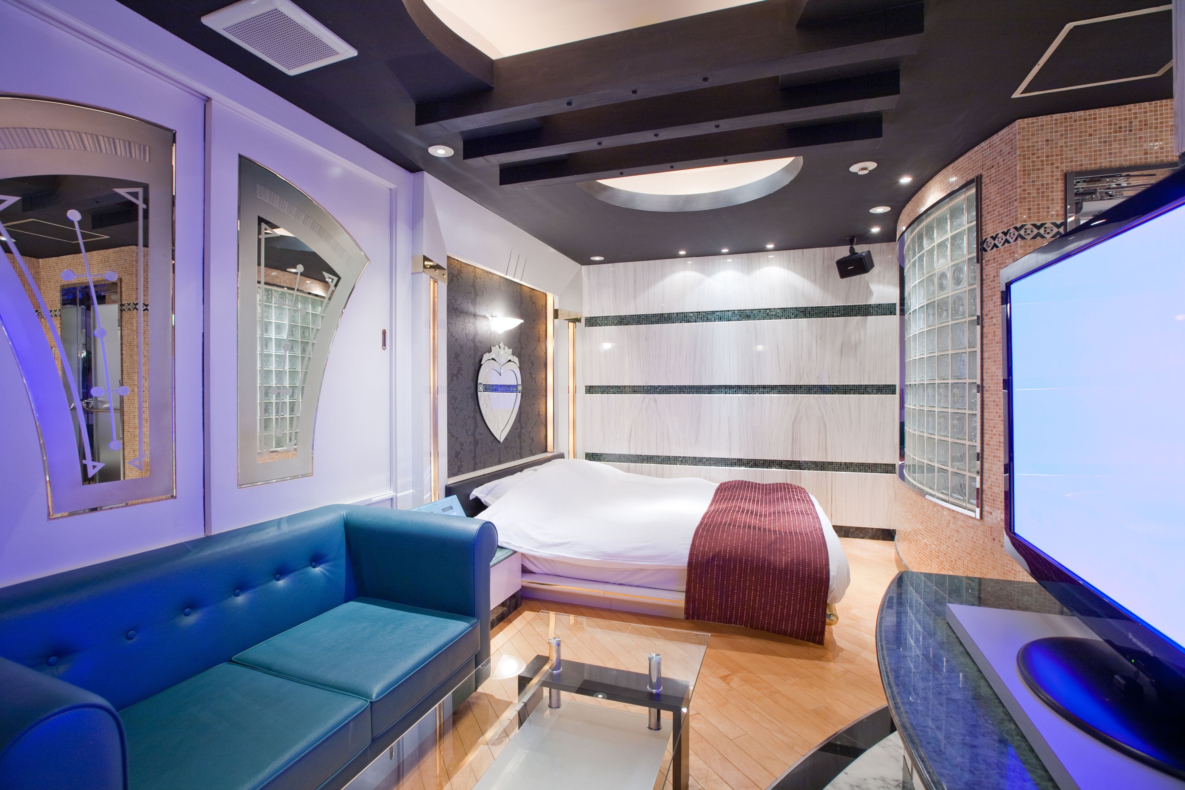 A modern hotel room with a blue sofa, a bed with a red blanket, a large TV, and a glass coffee table.