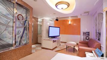Habitación de hotel con cama, televisor, zona de estar y baño visible a través de un separador de vidrio.