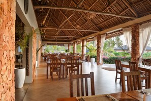 An outdoor dining area with wooden tables and chairs, surrounded by potted plants and a thatched roof structure.