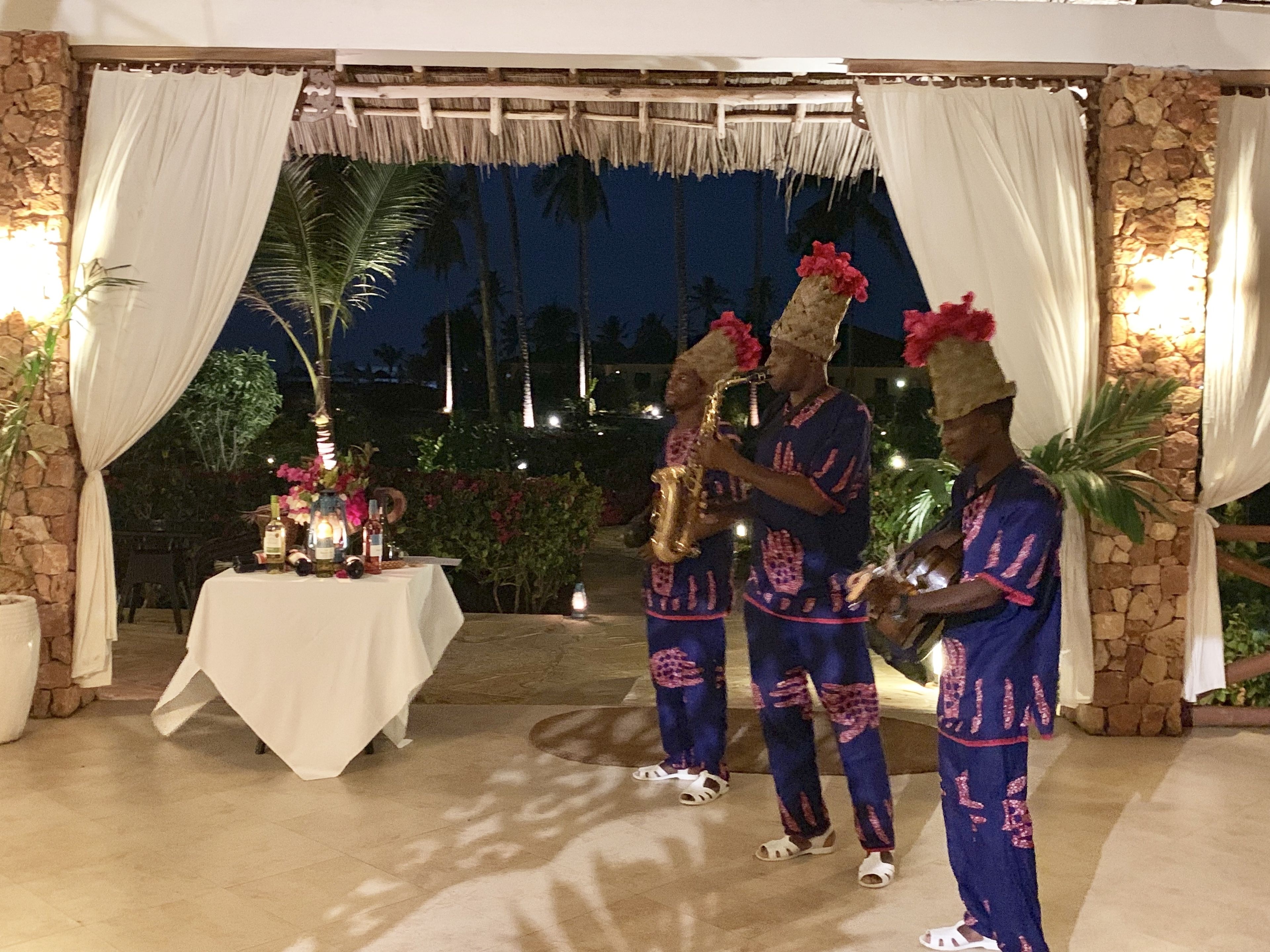 A stage with two musicians in traditional attire, a table with drinks and flowers, and a stone wall backdrop.