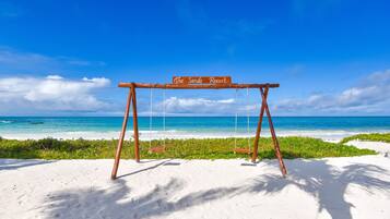 A wooden swing set with a sign reading "The Sands Resort" on a sandy beach with clear blue water and a clear sky.