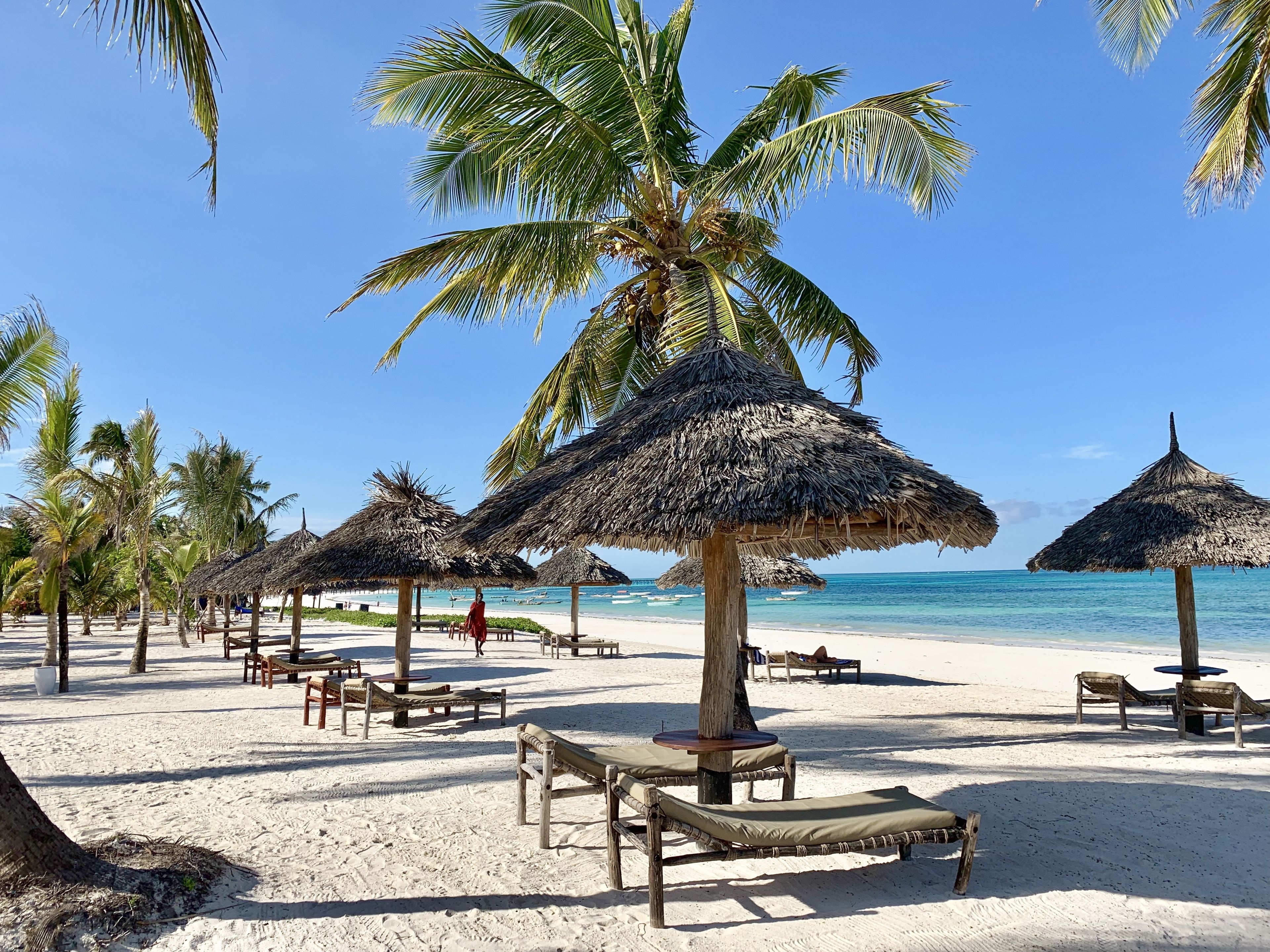 A beach with thatched umbrellas, wooden lounge chairs, and palm trees.