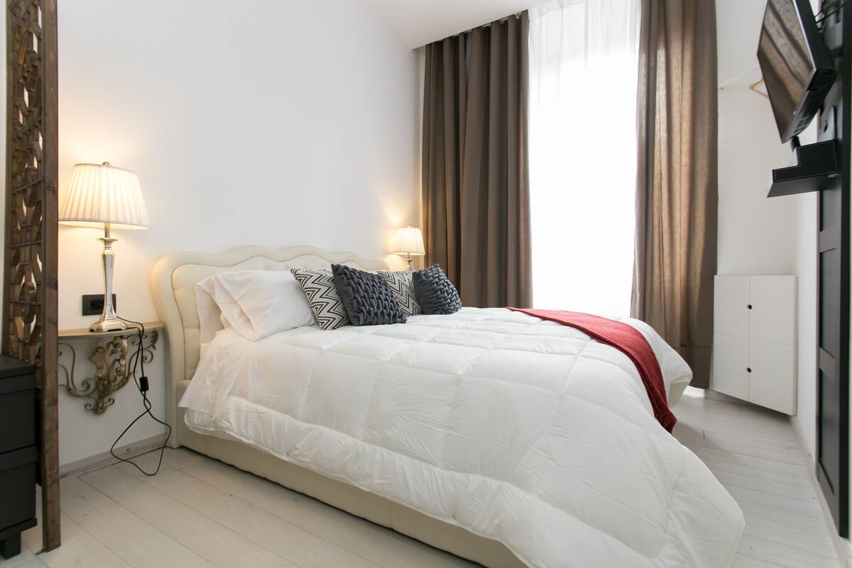 A neatly made bed with a white comforter and decorative pillows, a bedside lamp, and a wardrobe in a well-lit room.