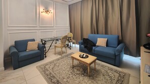 A living room with two blue sofas, a glass coffee table, a patterned rug, a chandelier, and a large window with curtains. - Aparthotel dei Mercanti (Milan)