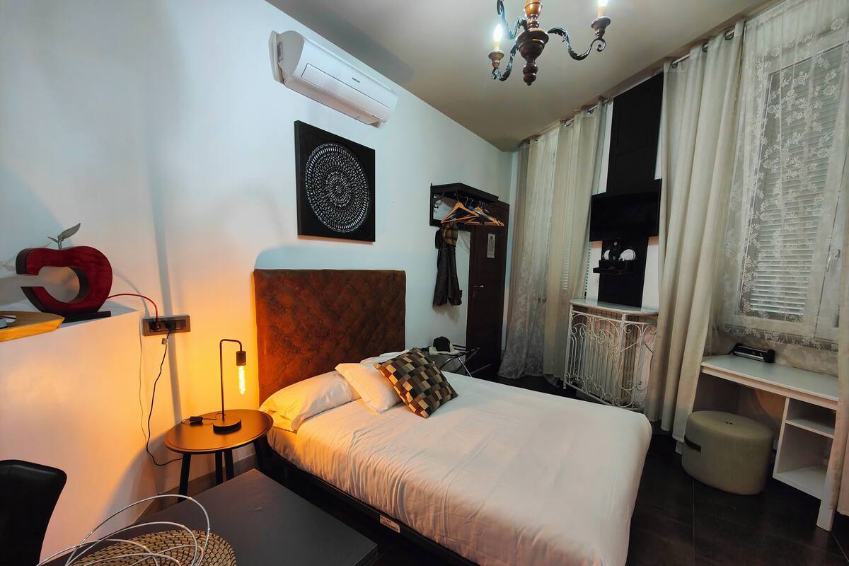 A hotel room with a bed, bedside table, lamp, air conditioning unit, and a chandelier.