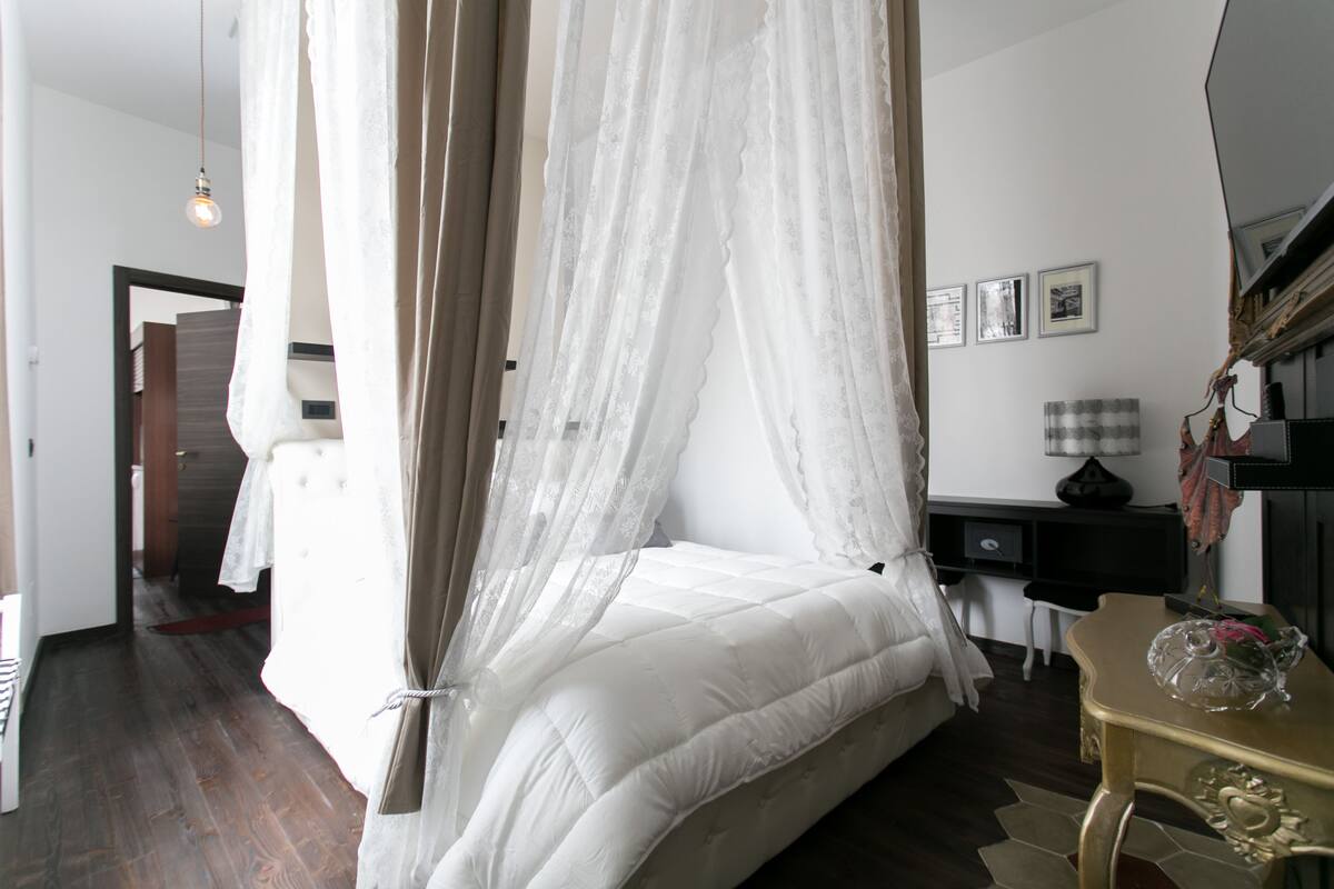 A bedroom with a canopy bed, a desk, a chair, and a small table with a decorative item.