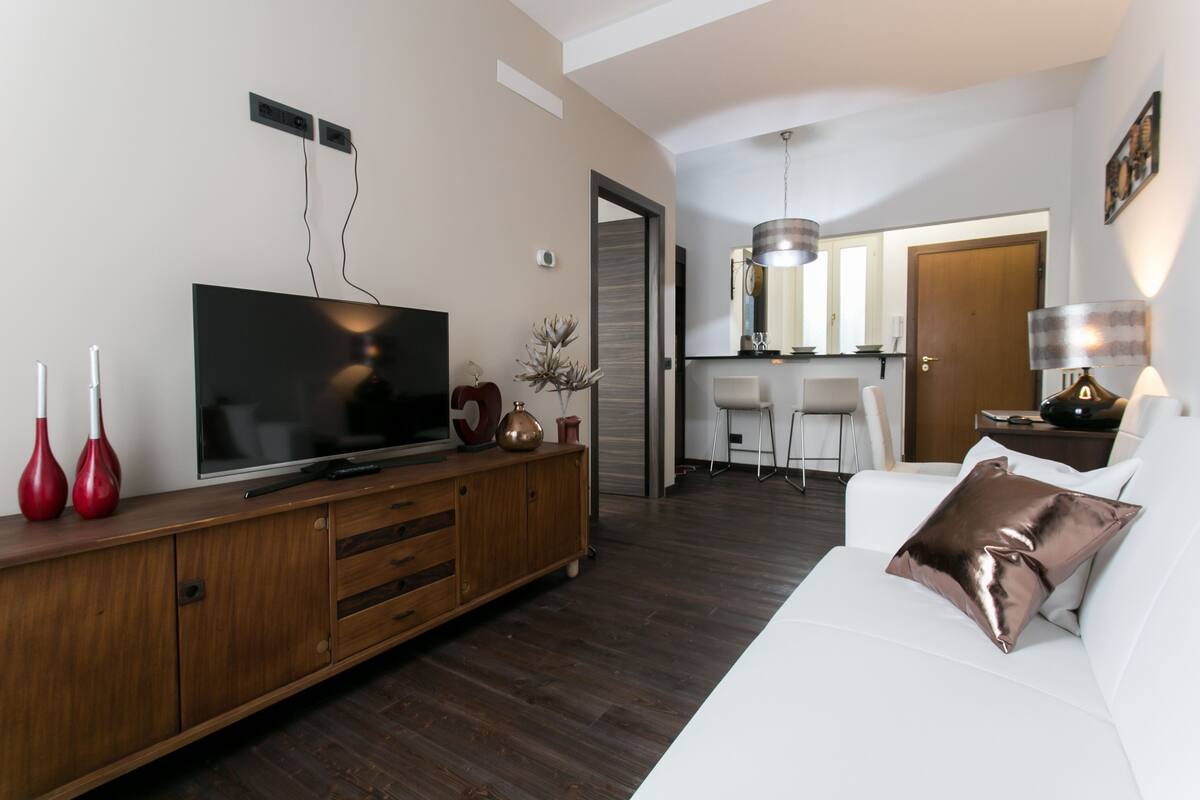 A modern living room with a flat-screen TV, wooden entertainment unit, white sofa, and a dining area in the background.