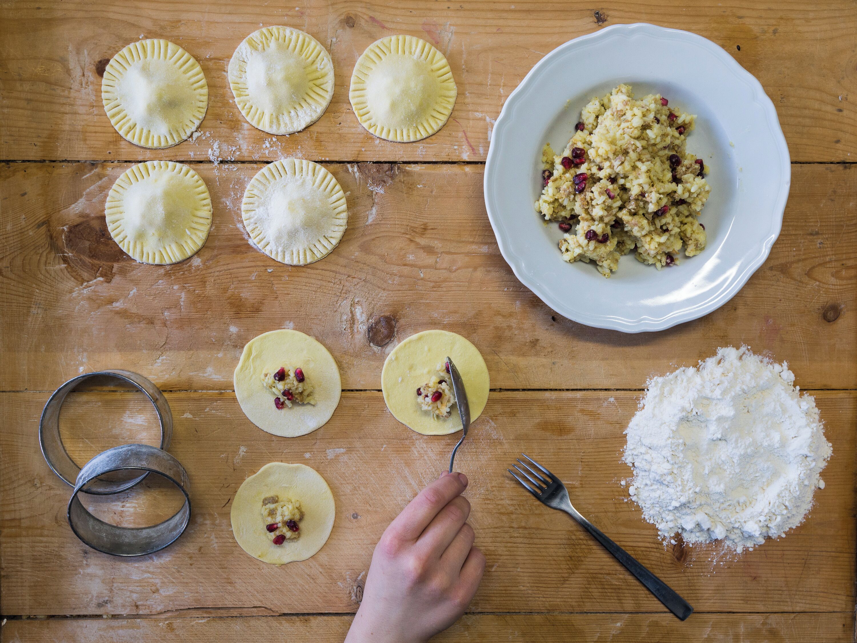 A wooden table with dough for pastries, a plate of mixed fruit, a rolling pin, and a metal cookie cutter.