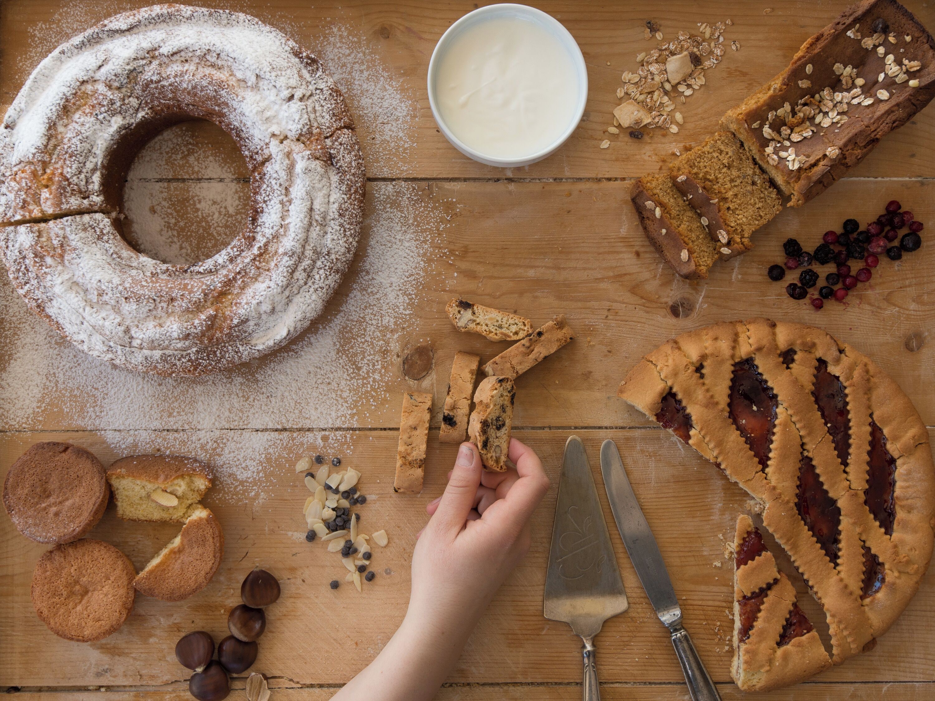 Une table en bois sur laquelle sont disposés divers produits de boulangerie, notamment un gâteau en forme de couronne, des biscuits et une tarte, ainsi qu’un bol de yaourt et un couteau.