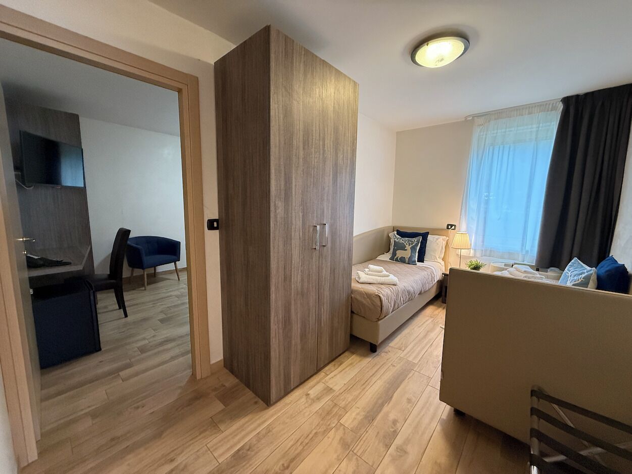 Una habitación de hotel moderna con un armario de madera, una cama con cubrecama blanco, un escritorio con silla y una ventana con cortinas.