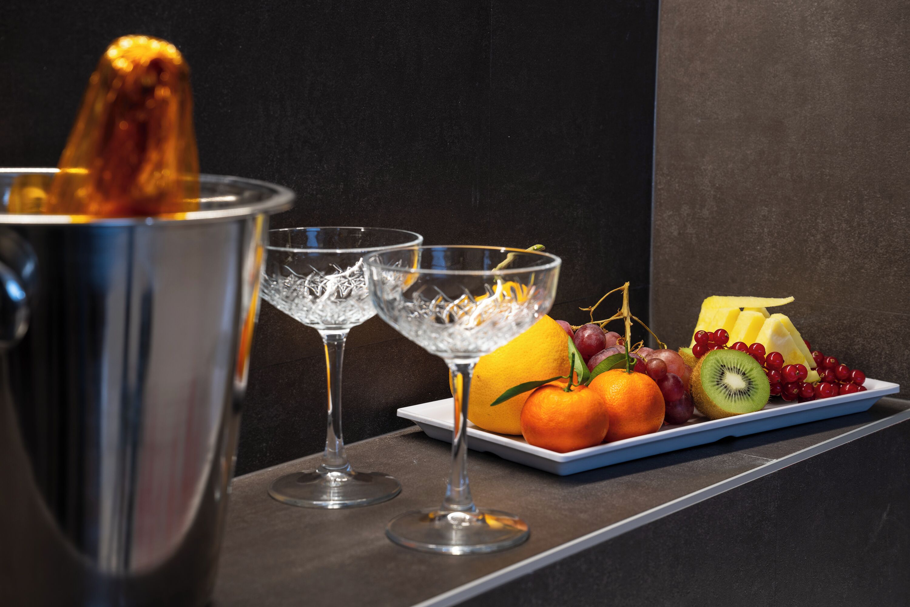 Una bandeja de frutas con uvas, naranjas, kiwi y un limón sobre un plato blanco, acompañada de dos vasos de agua y un recipiente metálico.