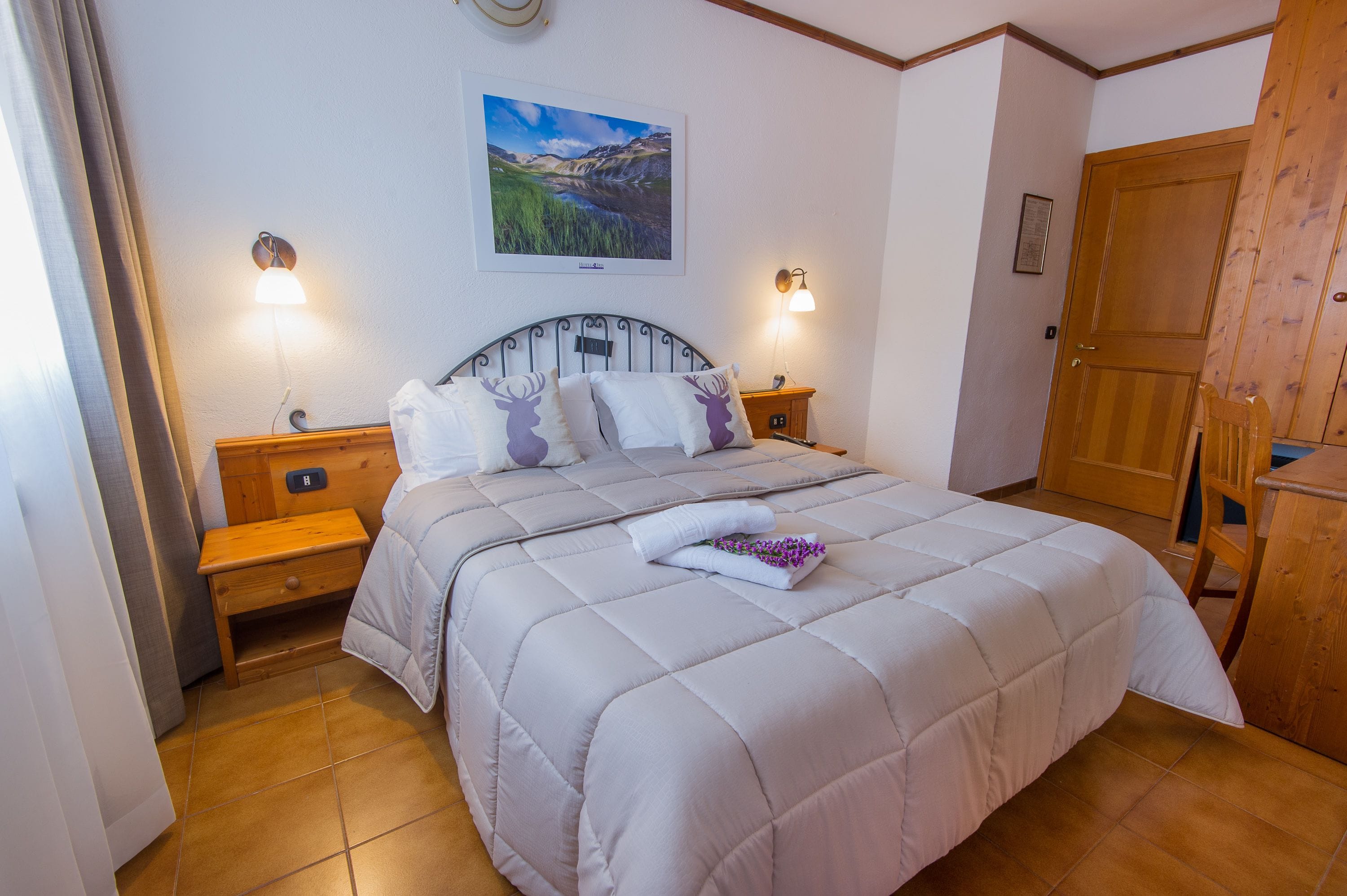 Una cama bien hecha con cabecera, dos mesitas de noche con lámparas, una puerta de madera y un cuadro en la pared.