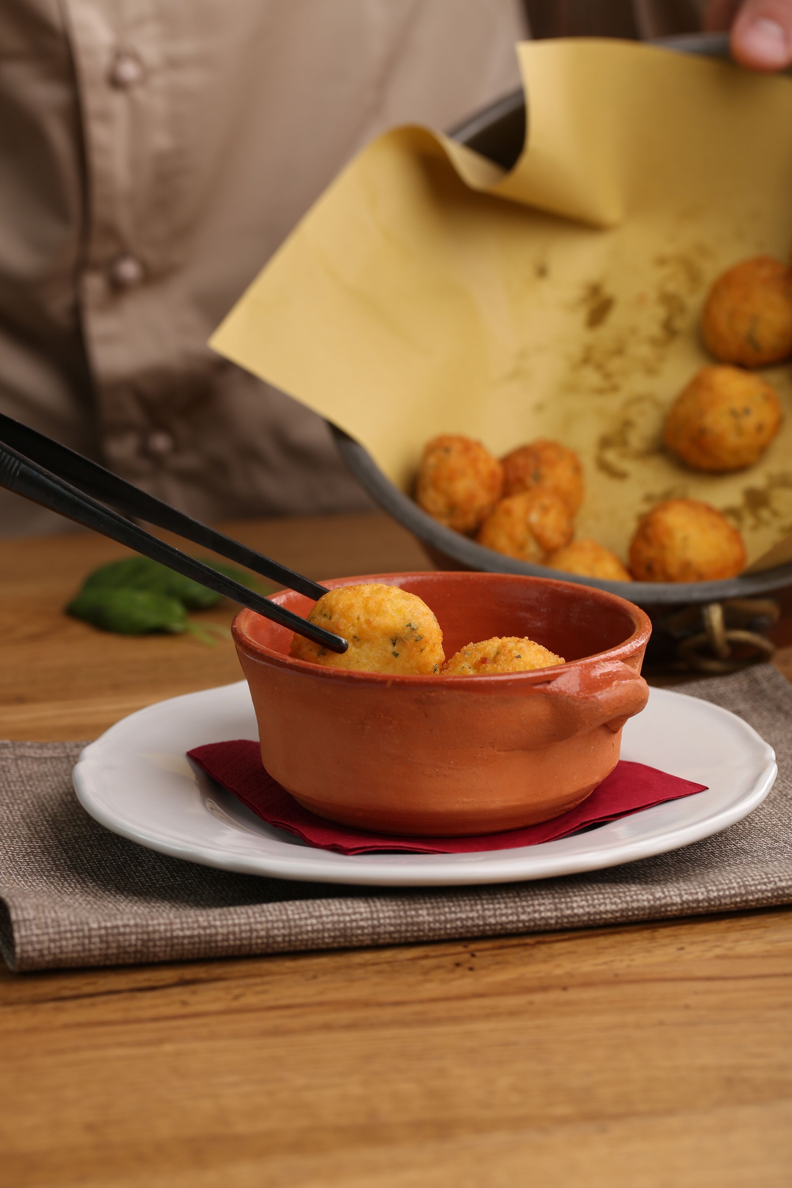Una pentola di terracotta con cibo su un tavolo, un tovagliolo rosso e un vassoio di metallo con altro cibo sullo sfondo.