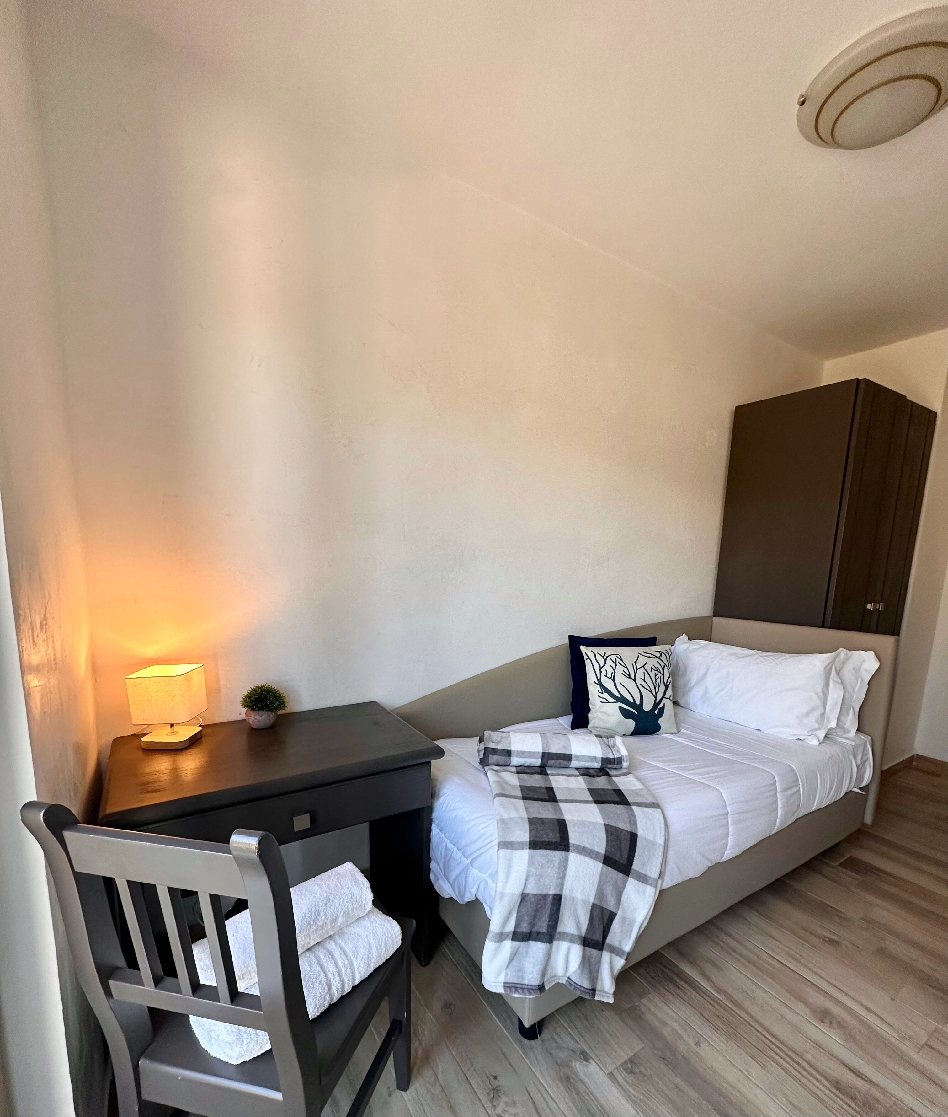 Een kleine, verzorgde slaapkamer met een enkel bed, een nachtkastje met lamp, een stoel en een garderobe.