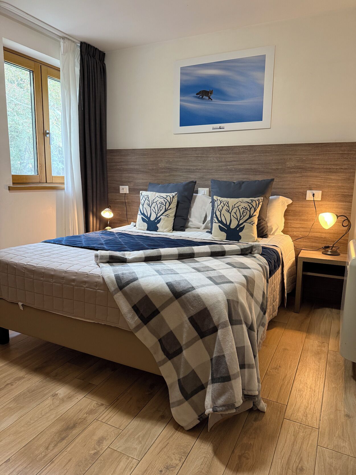 A neatly made bed with a checkered blanket, two pillows with tree designs, a nightstand with a lamp, a framed picture on the wall, and a window with curtains.