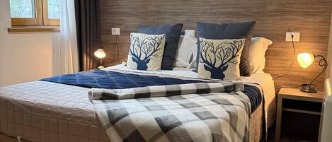 A neatly made bed with a checkered blanket, two pillows with tree designs, a nightstand with a lamp, a framed picture on the wall, and a window with curtains.