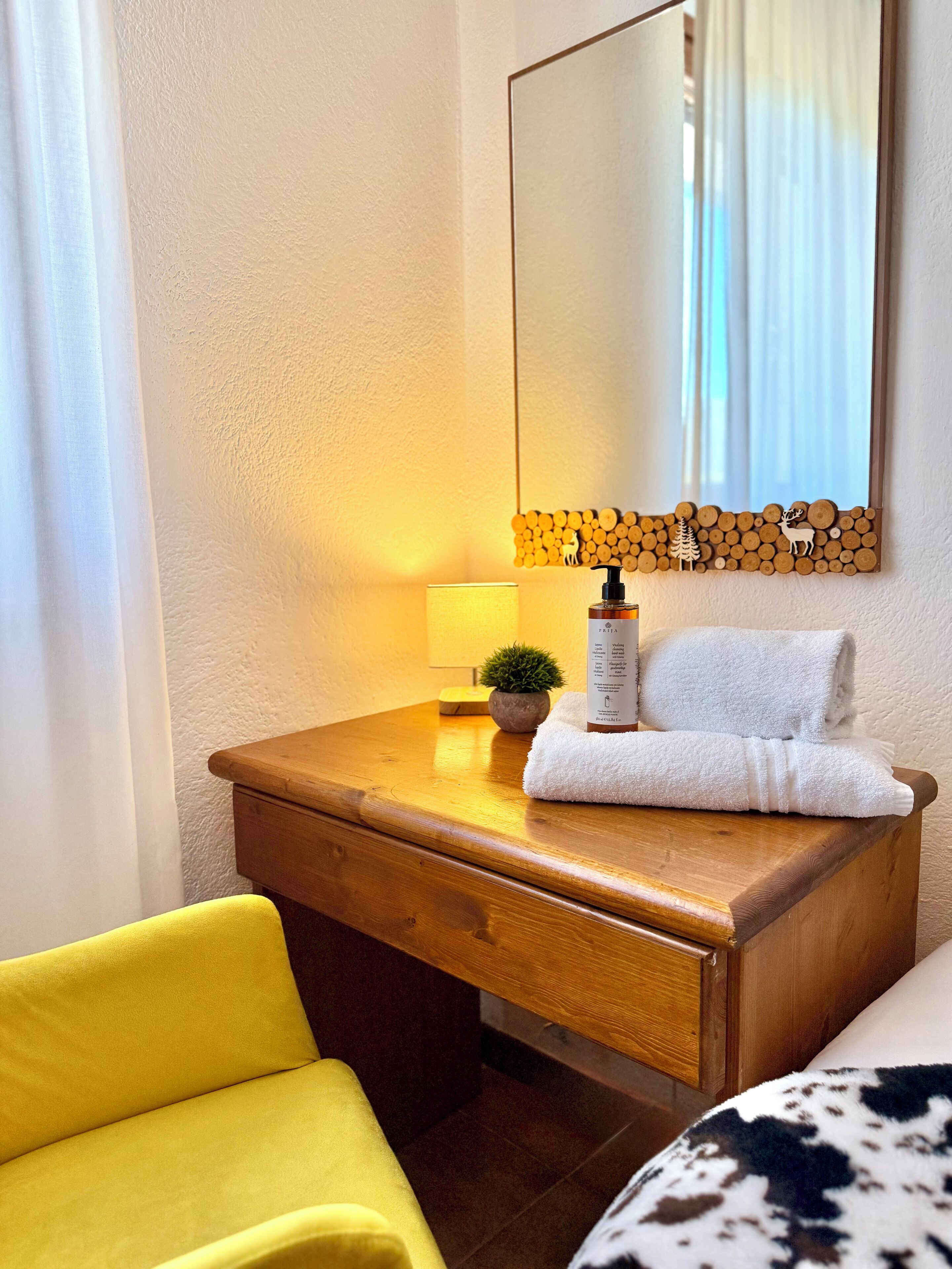 A hotel room with a wooden desk, a lamp, a bottle of lotion, a towel, and a mirror with a wooden log border.
