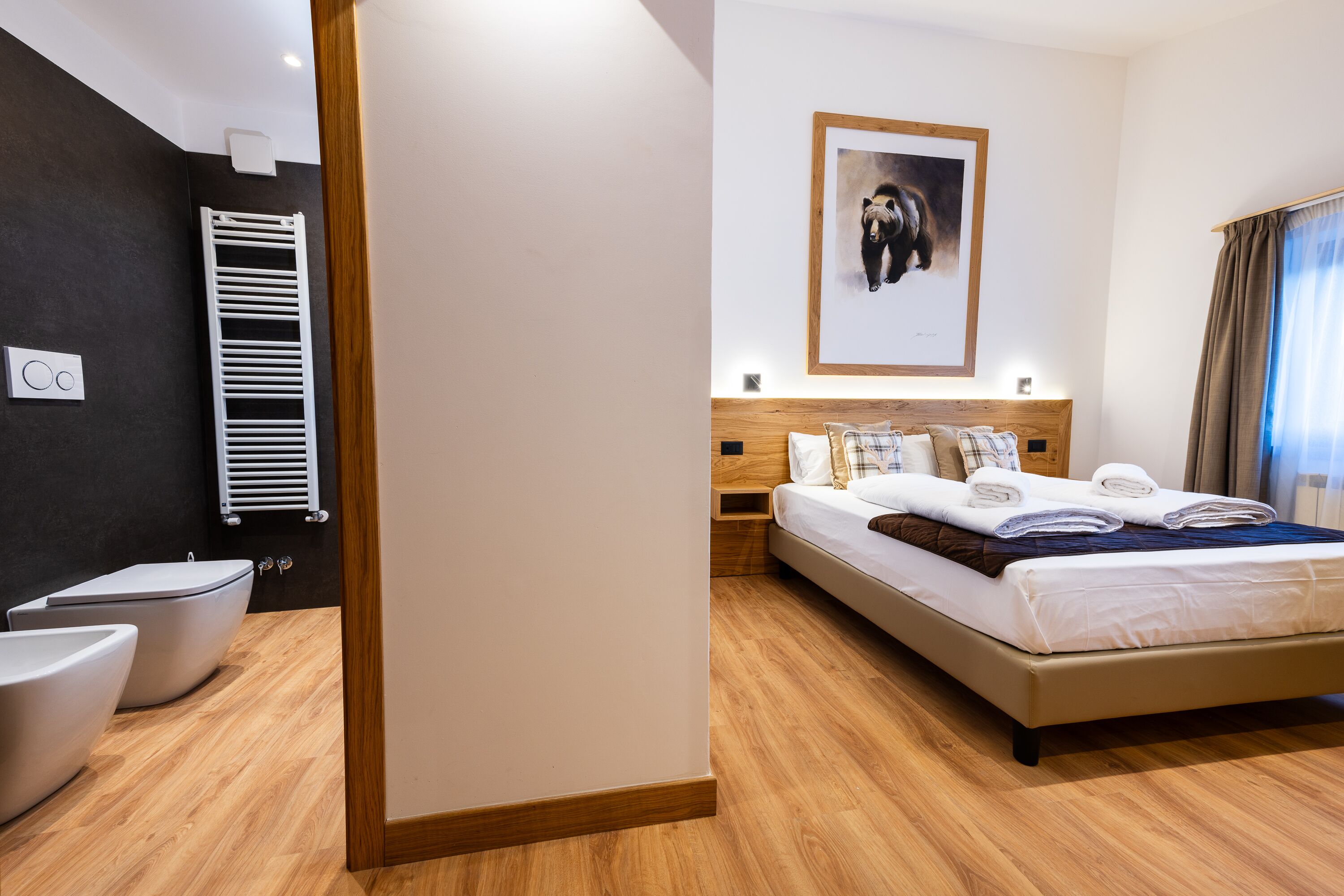 A modern hotel room with a wooden floor, a bed with white linens, a headboard with a framed picture, and a white radiator.