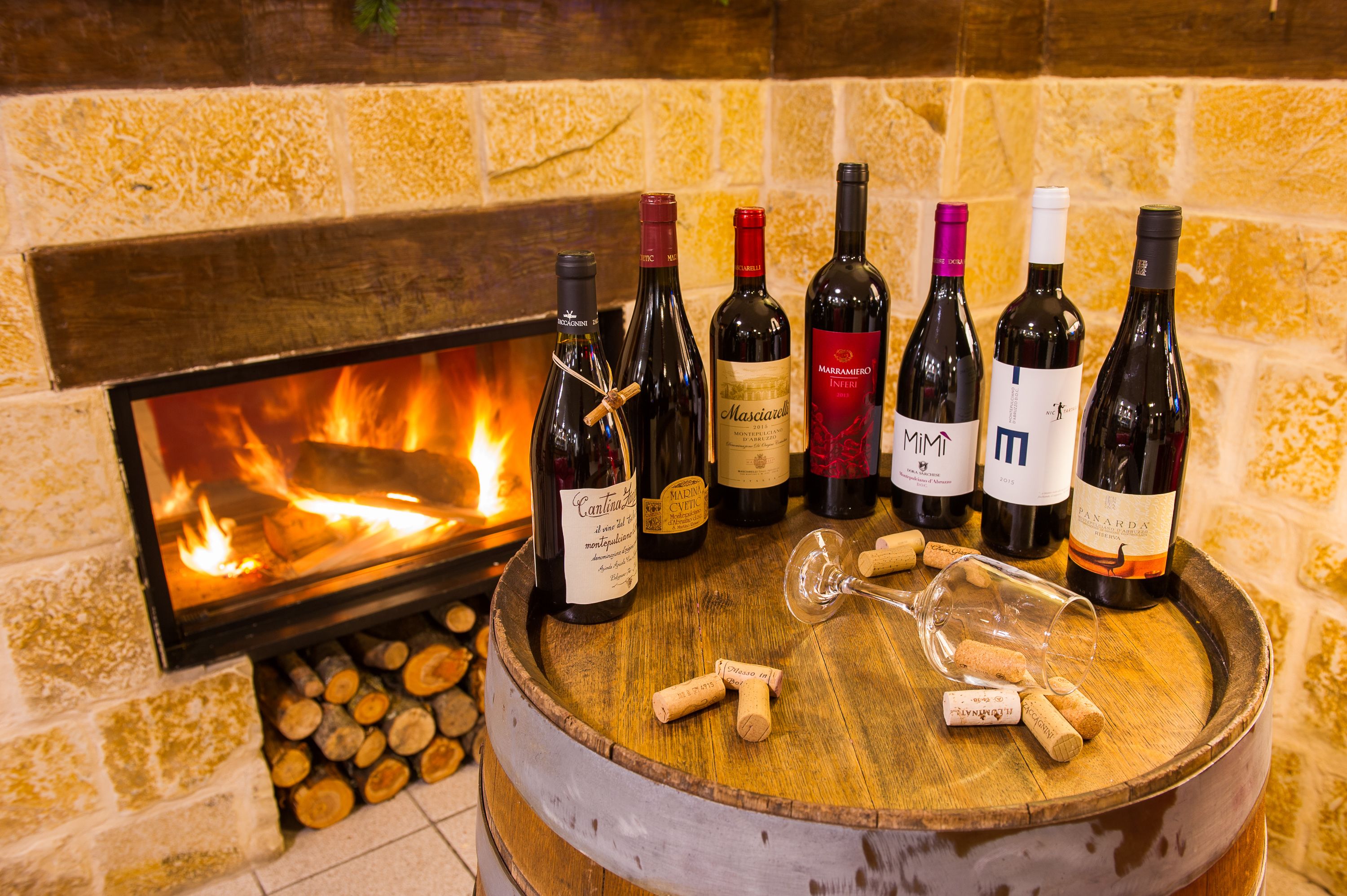 Un barril de madera con varias botellas de vino y tapones, situado frente a una chimenea con fuego encendido.