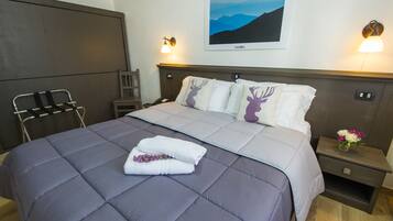 Una cama bien hecha con un edredón gris, dos almohadas con un diseño de ciervo y una toalla blanca doblada con flores púrpuras. Se observa una mesita de noche con un florero y una lámpara de pared.
