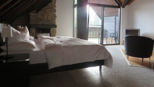 A bedroom with a stone fireplace, a bed with white linens, a black chair, and a balcony with a view. - Landgasthof Ralinger Hof (Ralingen)