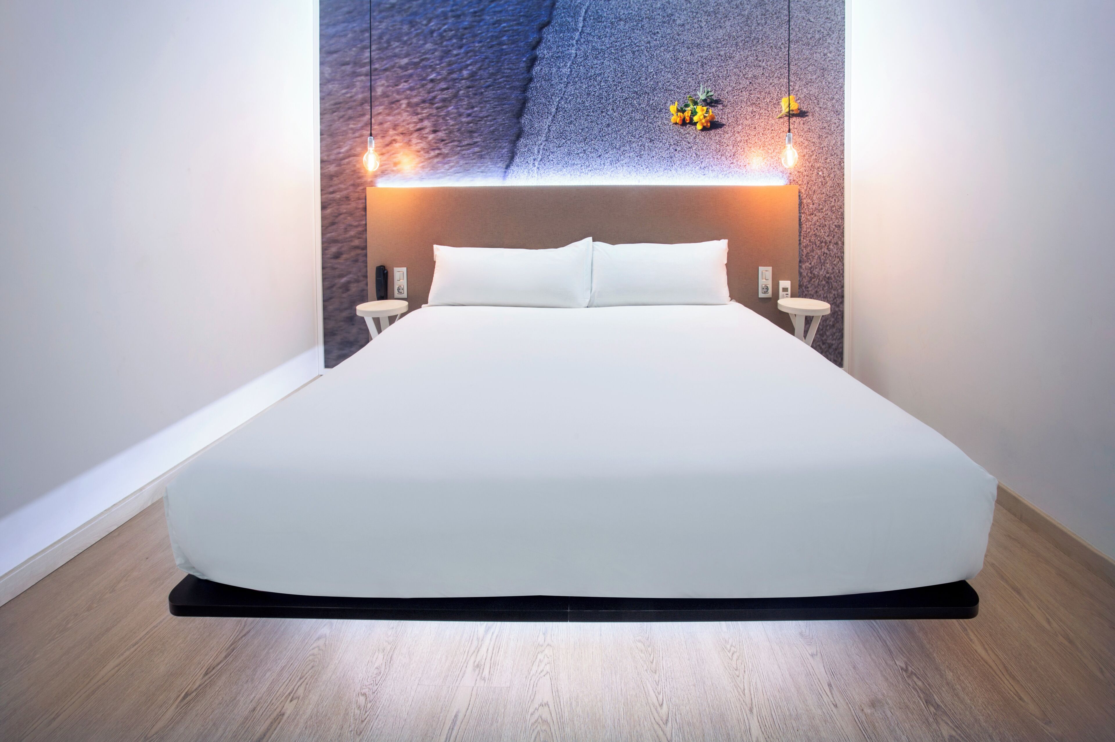 Ett modernt hotellrum med en stor säng, en sänggavel med inbyggd belysning, ett litet bord och en vägghylla med dekorativa föremål.