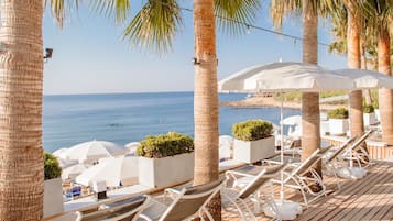 Une terrasse en bois, dotée de chaises longues et de parasols, qui offre une vue sur une plage bordée de palmiers et sur un ciel bleu azur.